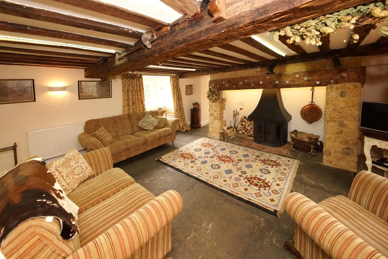 Salon de campagne aux poutres apparentes, avec cheminée en pierre, tapis et fauteuils à rayures.