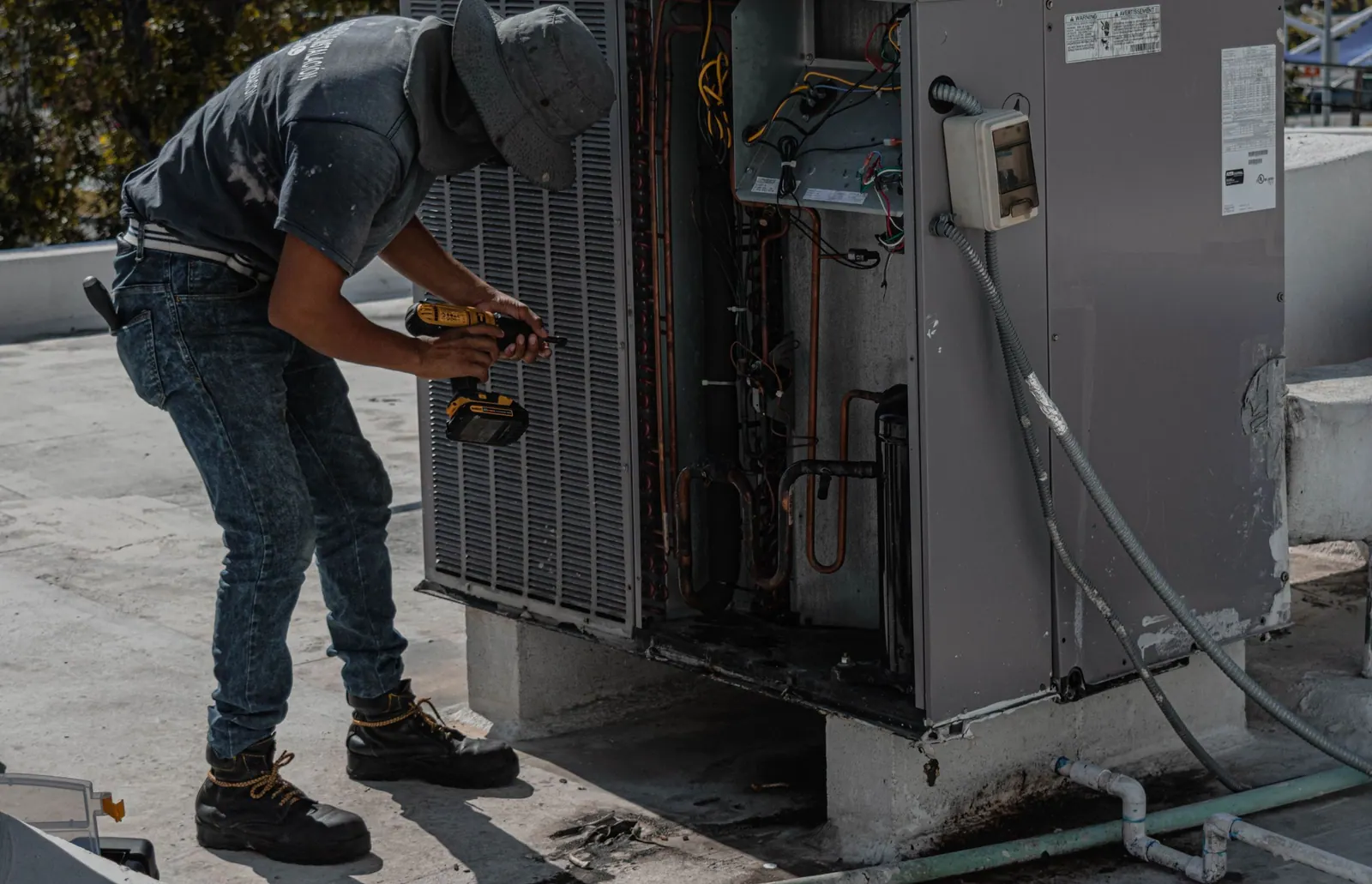 Technicien en chapeau qui démonte le capot d’une unité extérieure de climatisation sur un toit-terrasse.