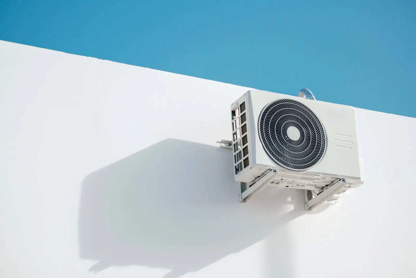 Unité extérieure de climatisation fixée sur un mur blanc, sous un ciel bleu.