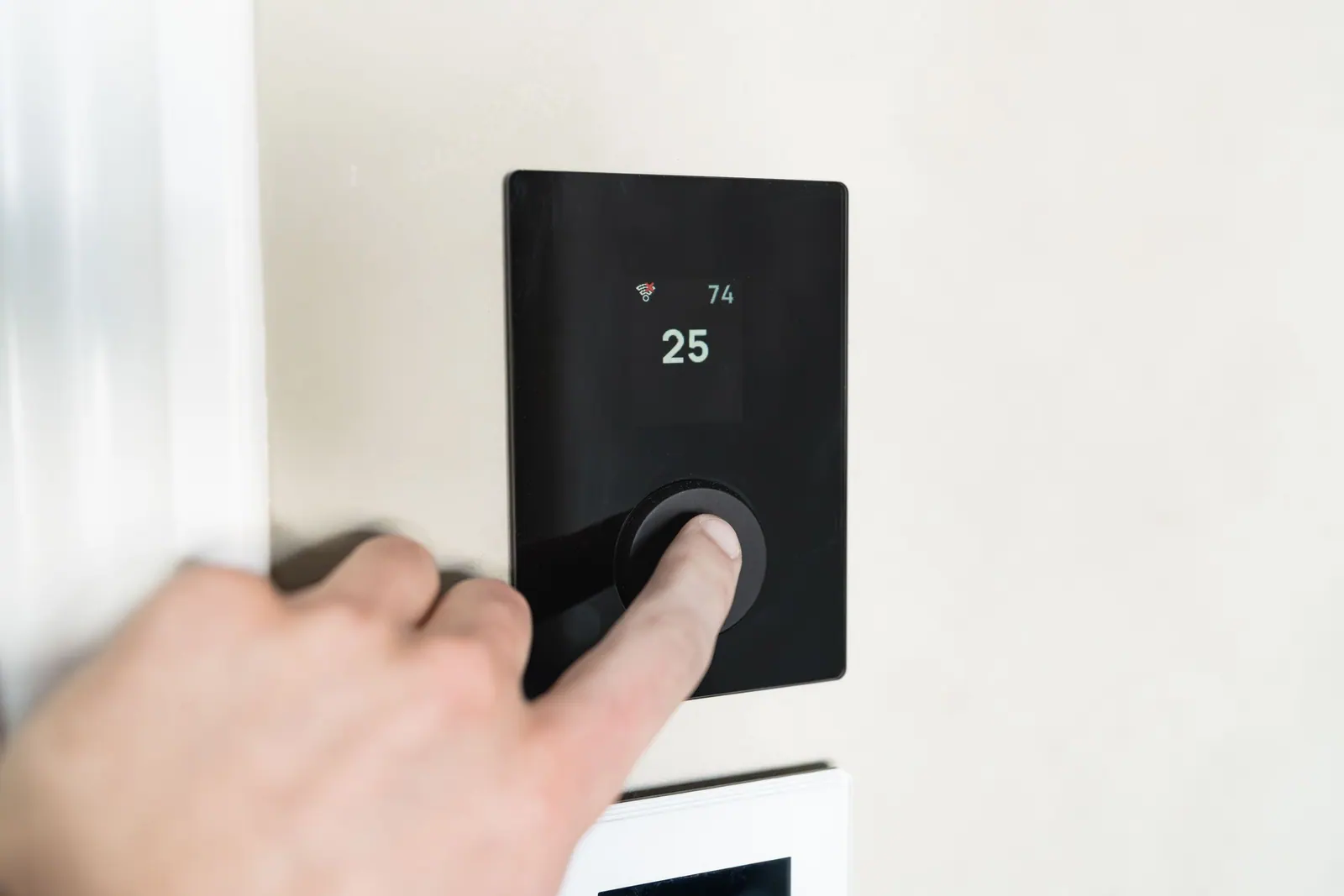 Doigt qui règle un thermostat mural noir affichant 25 degrés.
