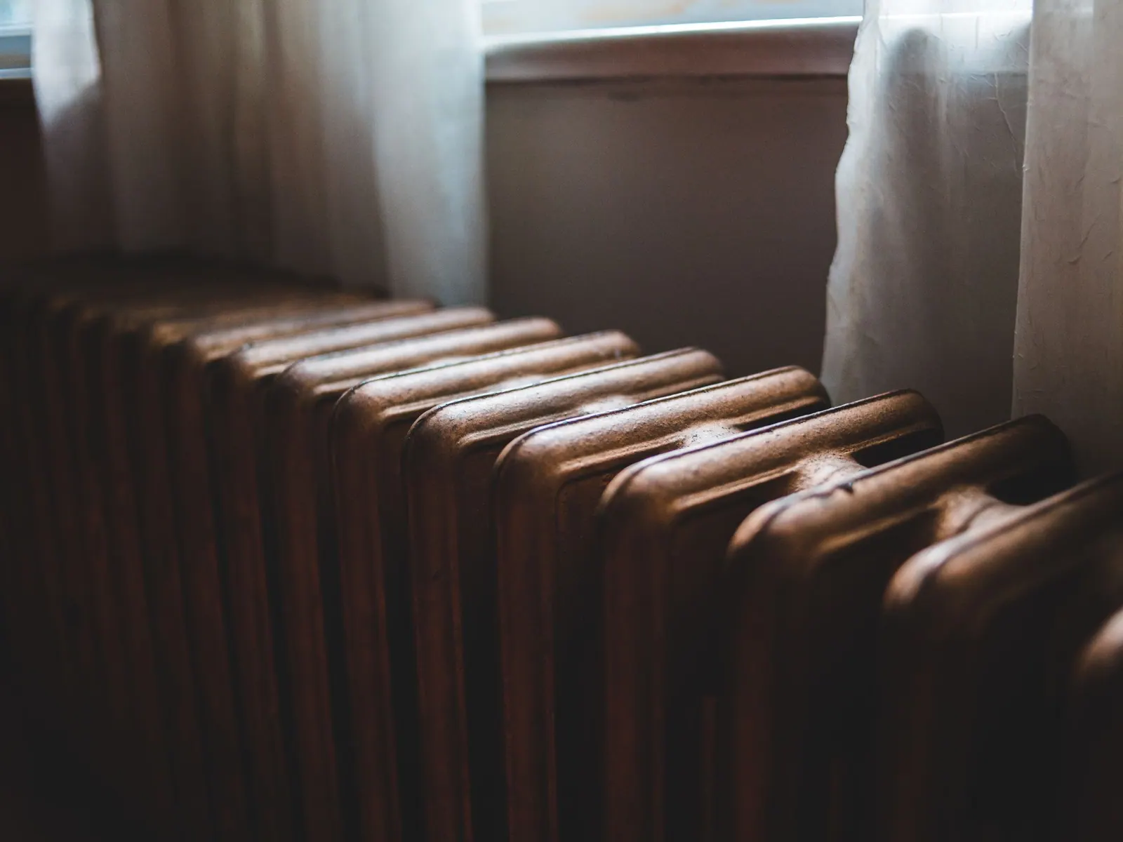 Vieux radiateur en fonte brun foncé posé sous une fenêtre aux rideaux clairs.