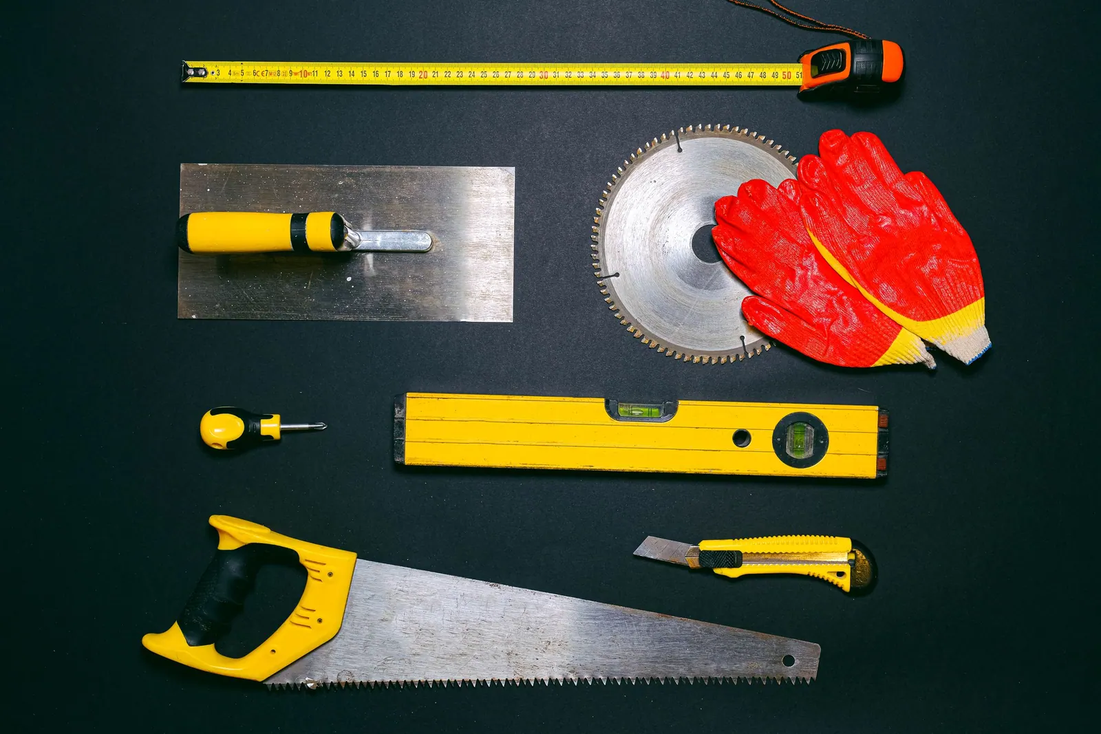 Outils de maçon posés à plat sur fond noir : platoir, niveau à bulle, scie, mètre ruban et gants.