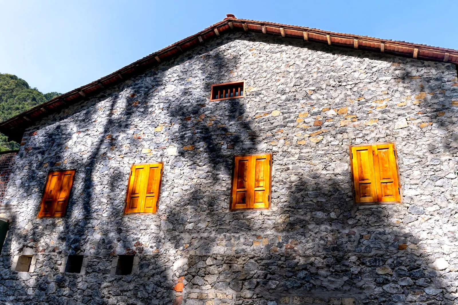 Façade en pierres apparentes sous un toit de tuiles, avec quatre volets en bois.