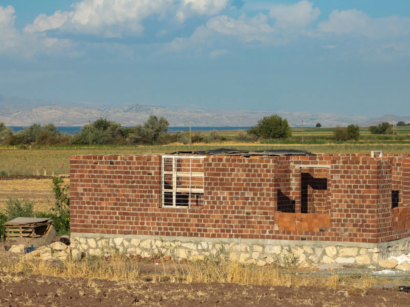 Gros œuvre en briques rouges d’une maison en construction, sur un soubassement en pierre, au milieu des champs.
