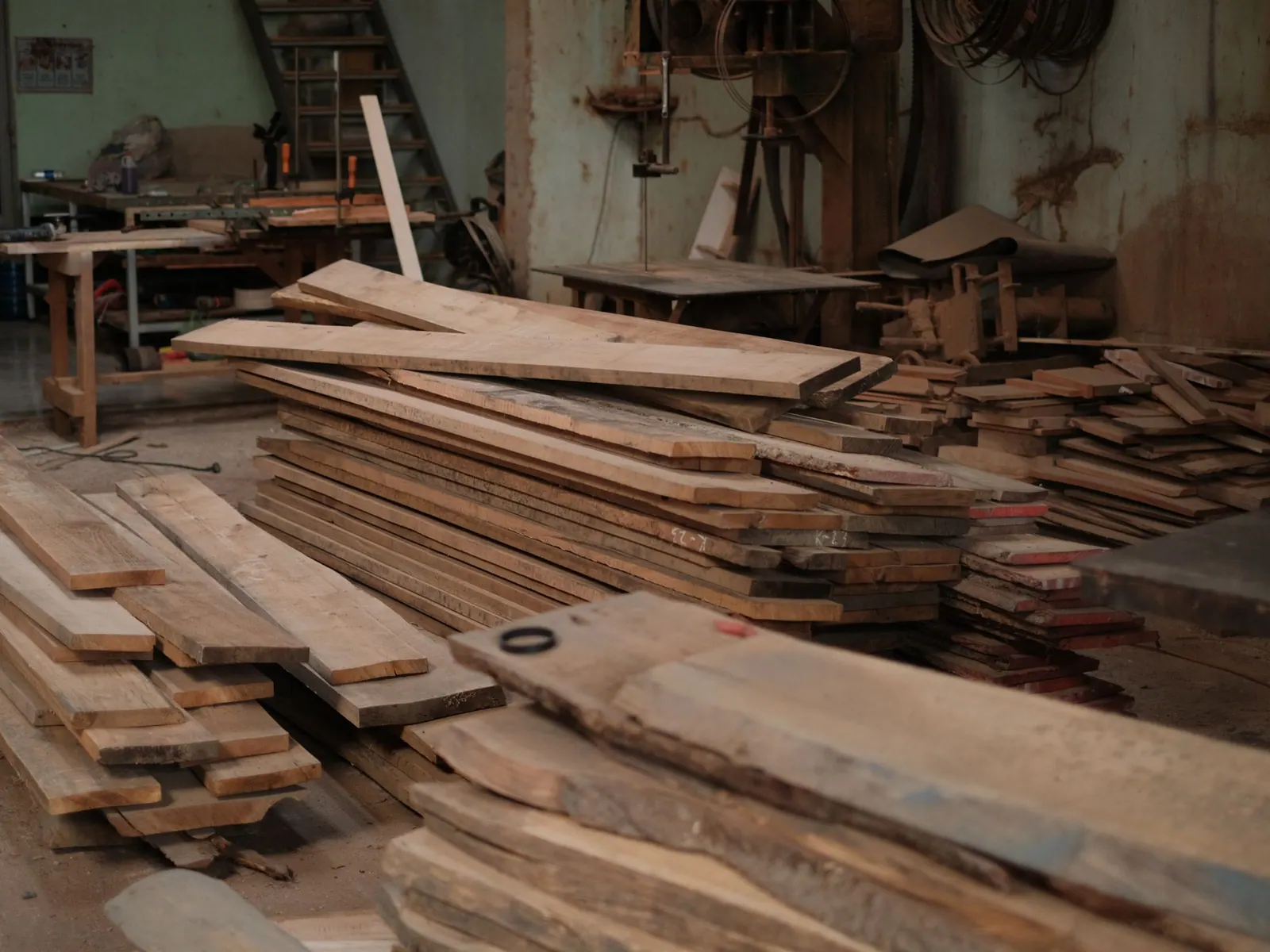Piles de planches de bois brut dans un atelier de menuiserie, avec une scie à ruban au fond.