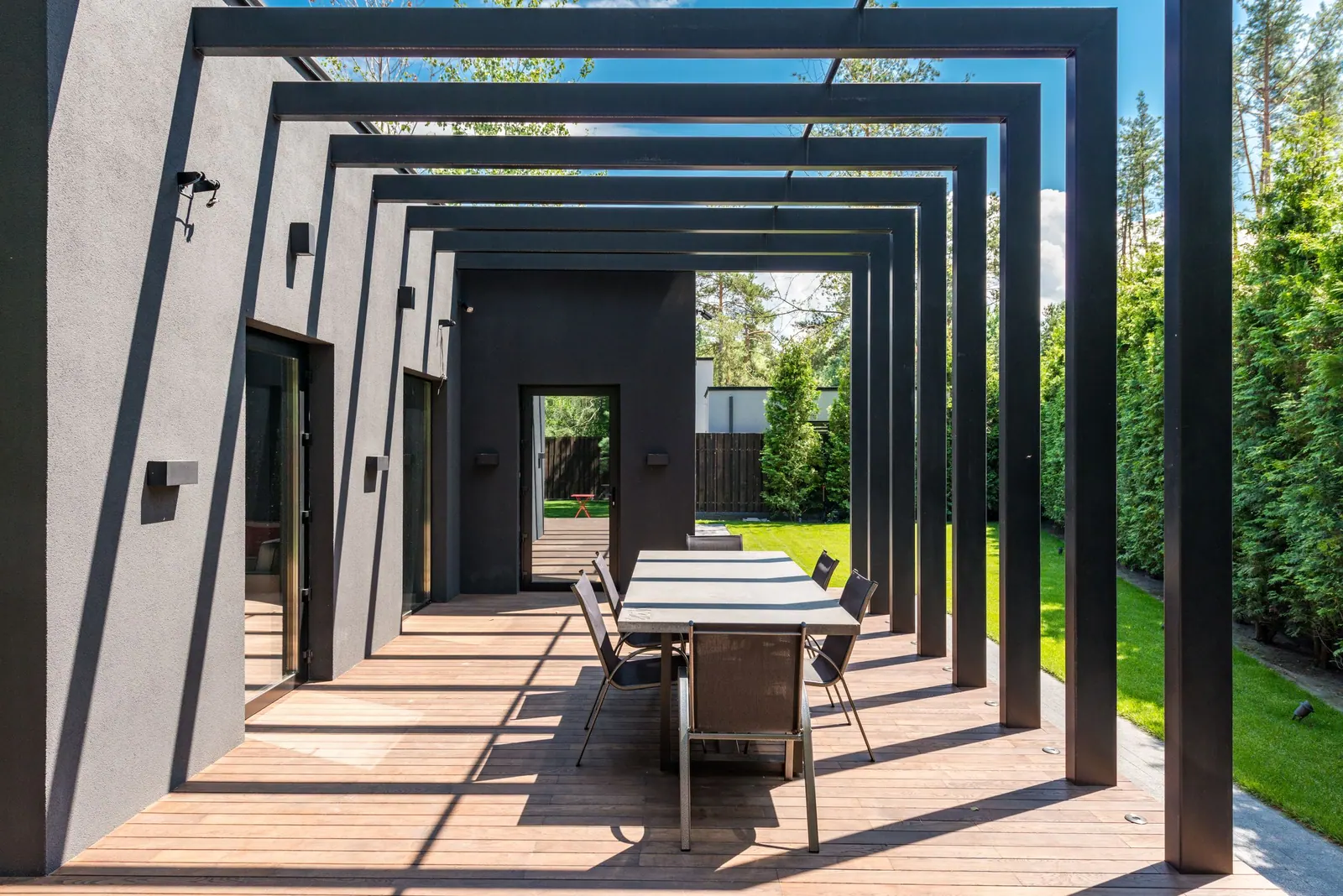 Terrasse en bois sous une pergola métallique noire, avec table et chaises ouvertes sur la pelouse.