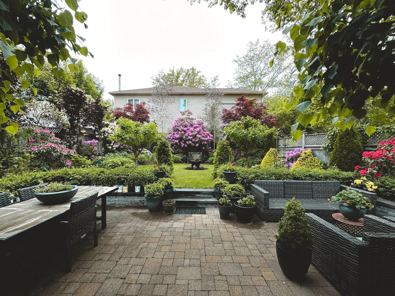 Jardin aménagé avec terrasse en pavés, massifs fleuris, buis taillés et salon de jardin.