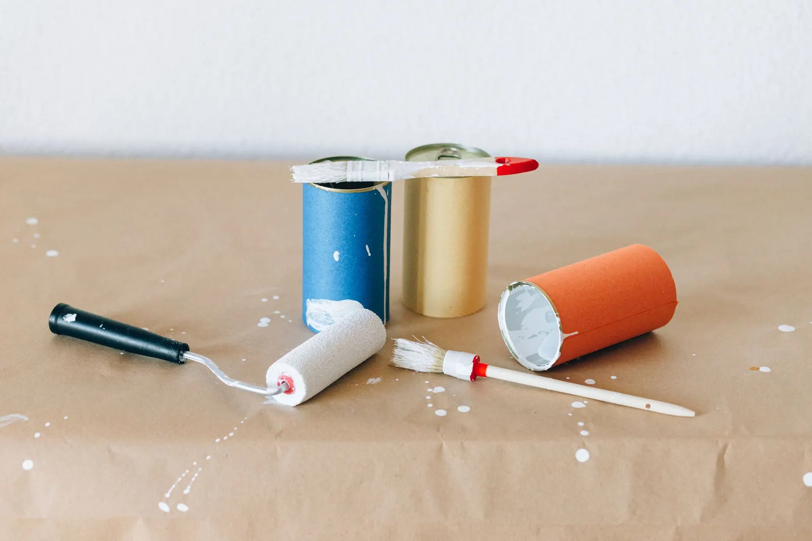 Pots de peinture, rouleau et pinceau posés sur du papier kraft éclaboussé de blanc.