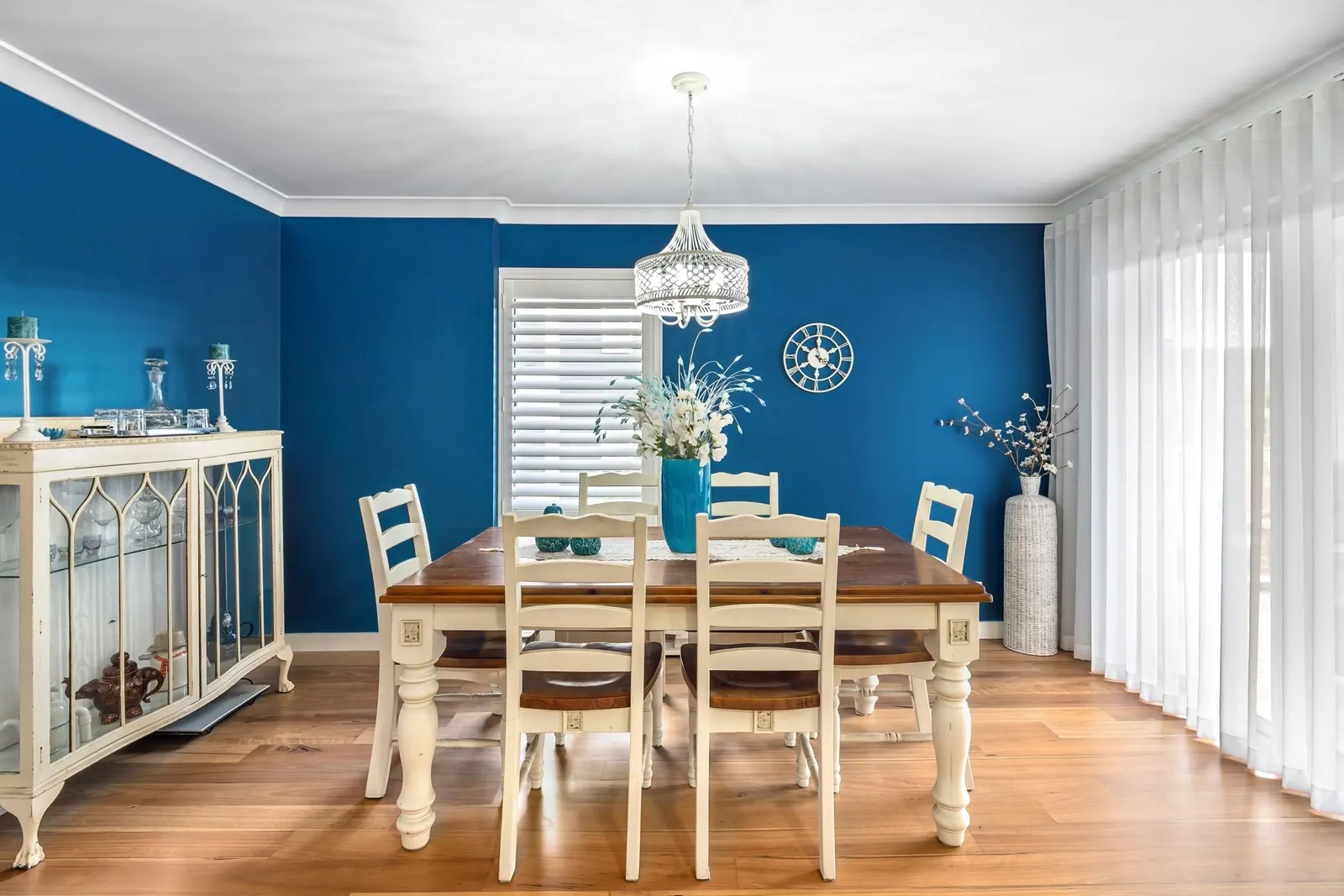 Salle à manger avec un mur d’accent bleu profond, une table en bois, des chaises blanches et un lustre.