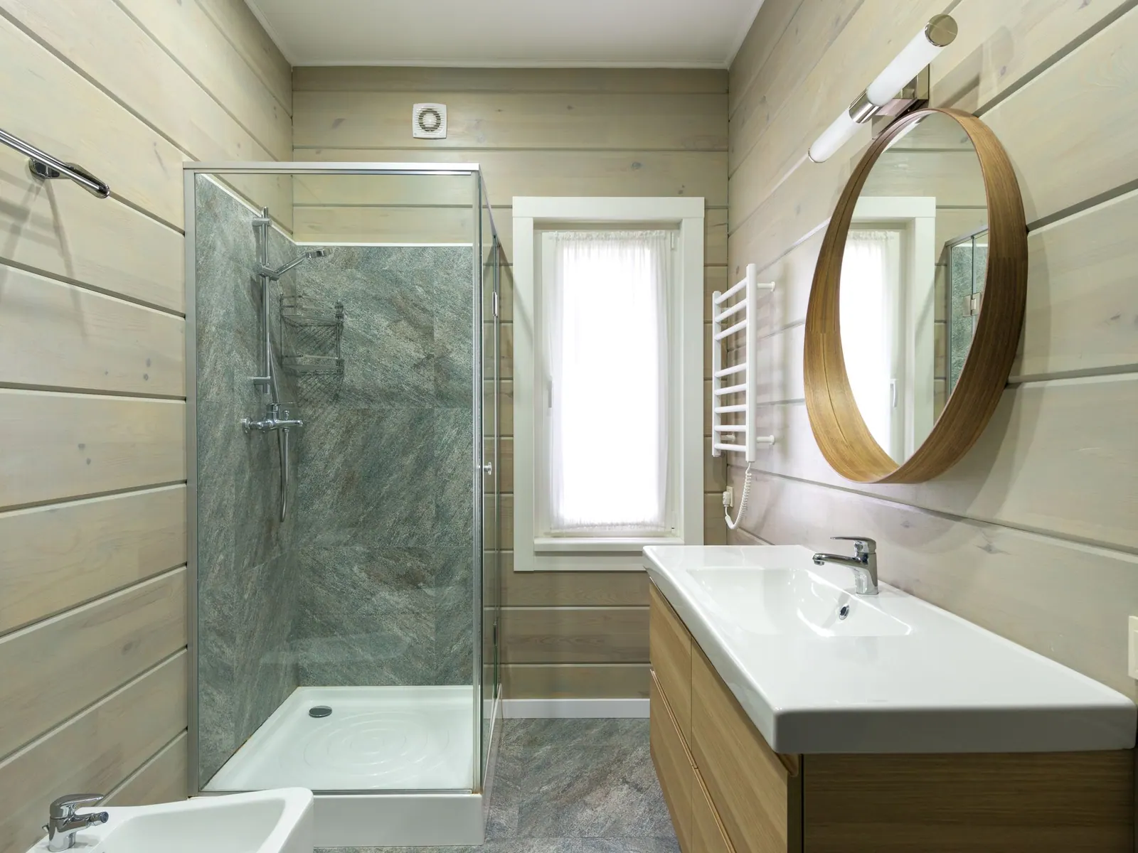 Salle de bain neuve avec douche vitrée, vasque sur meuble en bois, miroir rond et sèche-serviettes blanc.