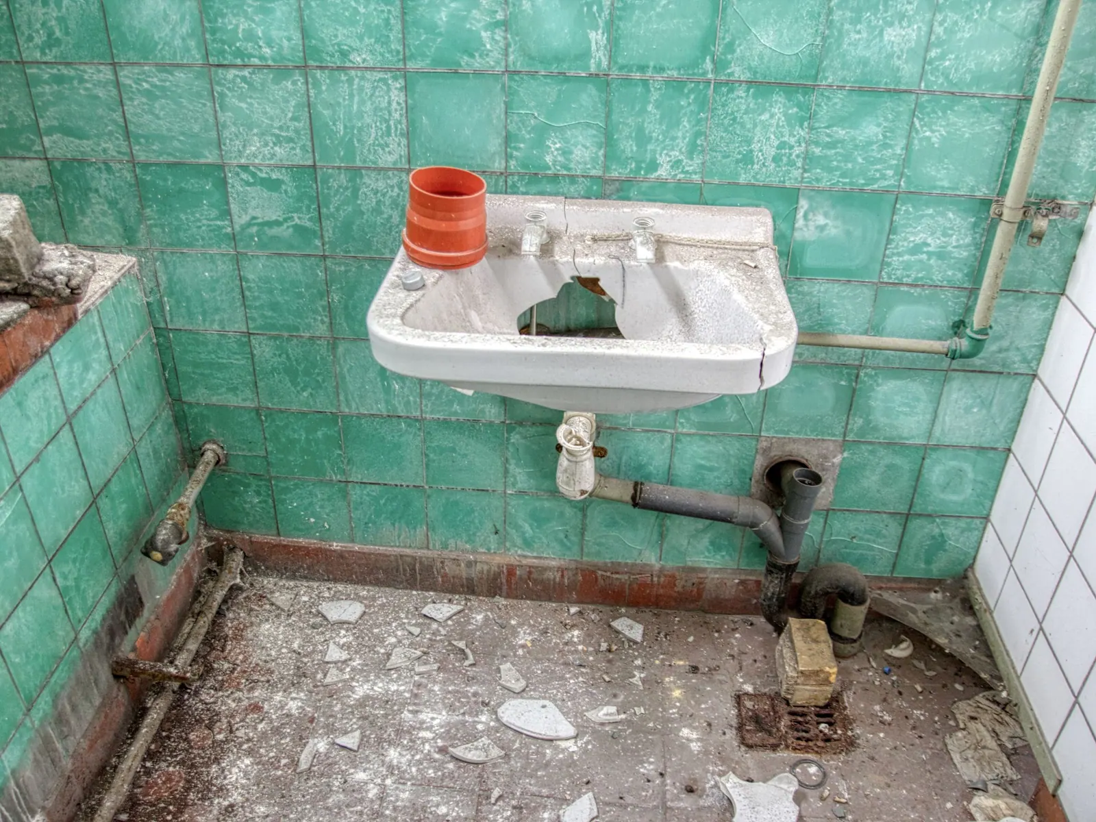 Salle d’eau abandonnée avec un lavabo cassé, des carreaux verts et des tuyaux rouillés.