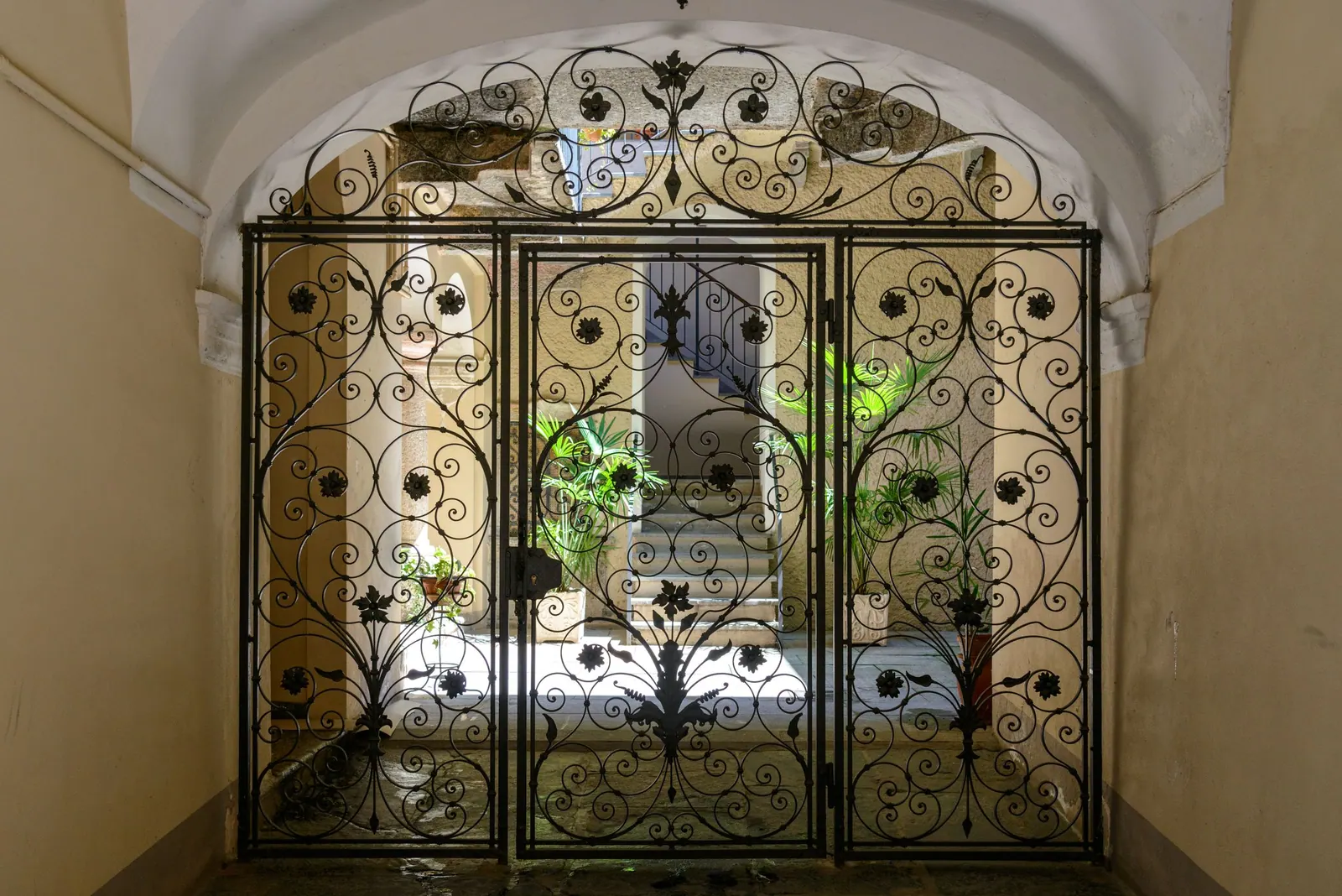 Portail en fer forgé à volutes et fleurs, fermant un passage voûté ouvert sur une cour.